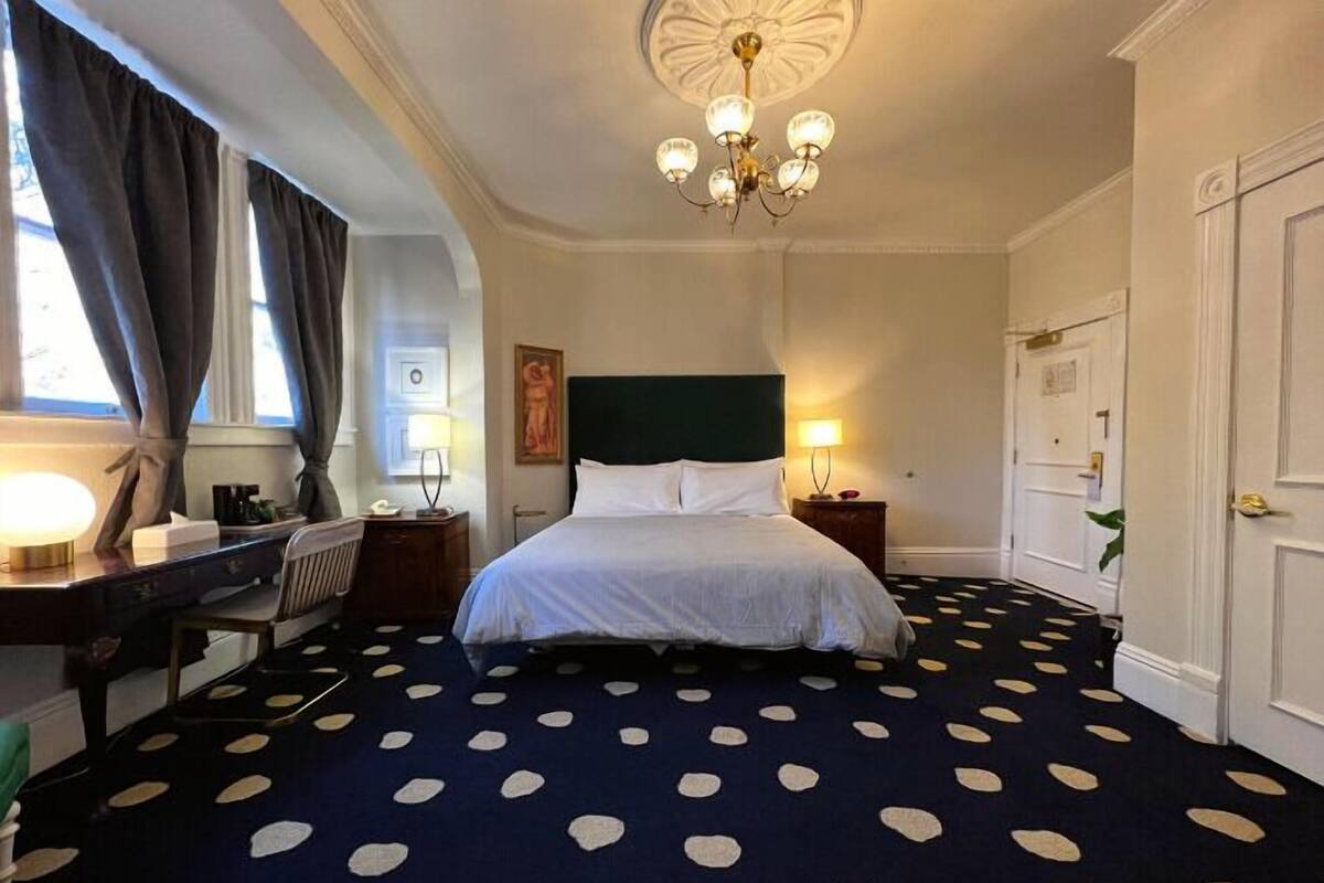 A hotel room with a bed, a desk, a chair, a lamp, a nightstand, a chandelier, and a door.