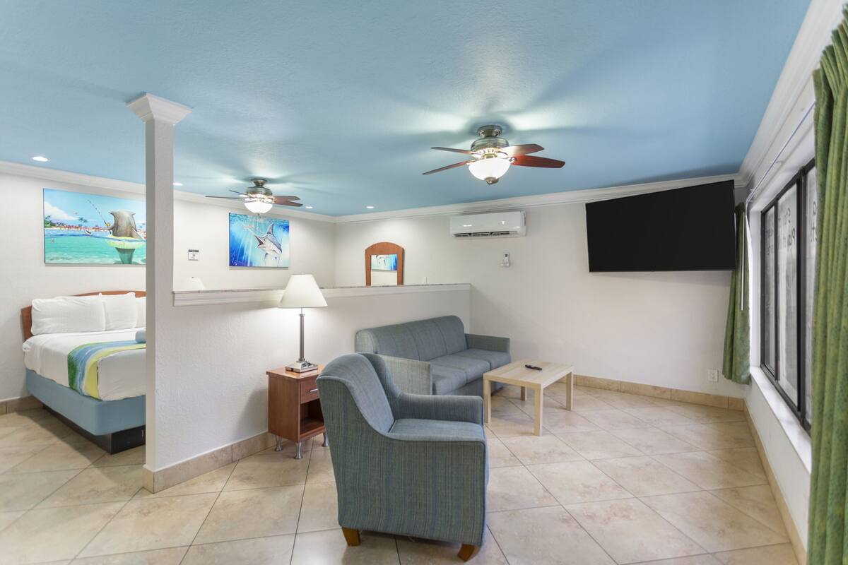 Ein Zimmer mit einem Bett, einem Sofa, einem kleinen Tisch, einer Lampe, einem Deckenventilator und einem an der Wand befestigten Fernseher.