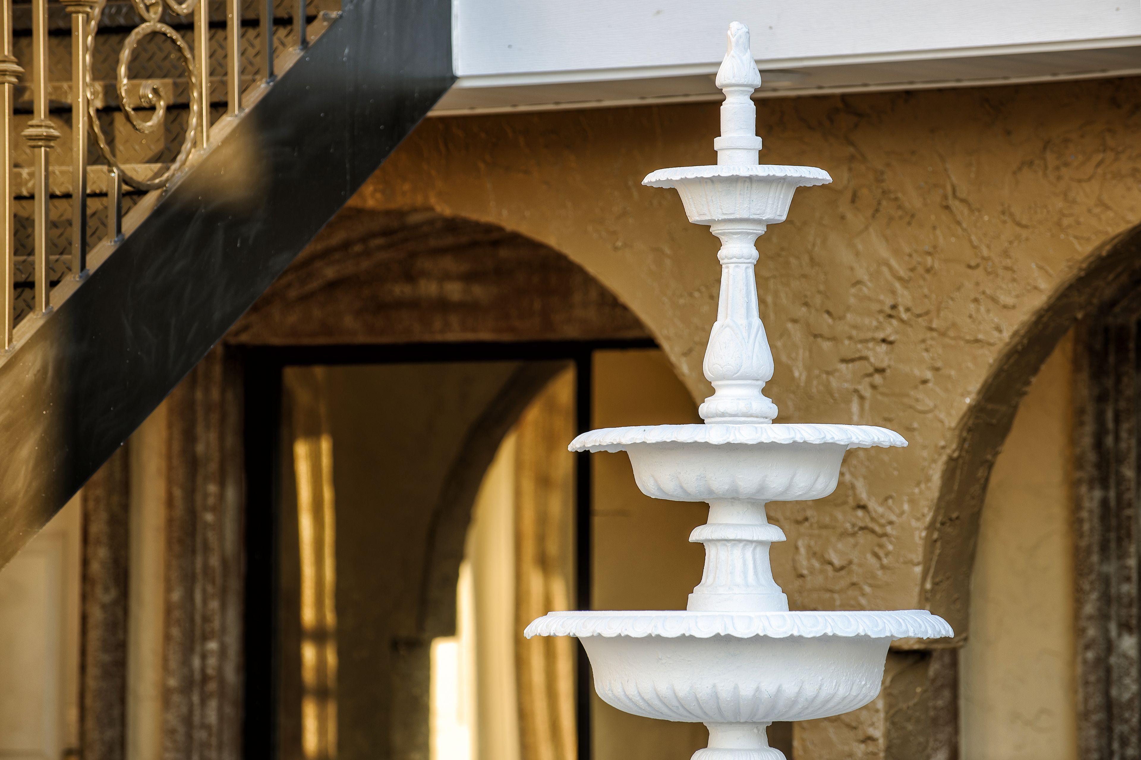A white, multi-tiered fountain in front of a building with a black staircase and arched doorways.