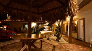 A thatched-roof outdoor area with a wooden table and benches, a fountain, and a staircase leading to an upper level.