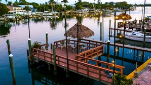 A wooden dock with chairs and a thatched umbrella, surrounded by calm water and boats.