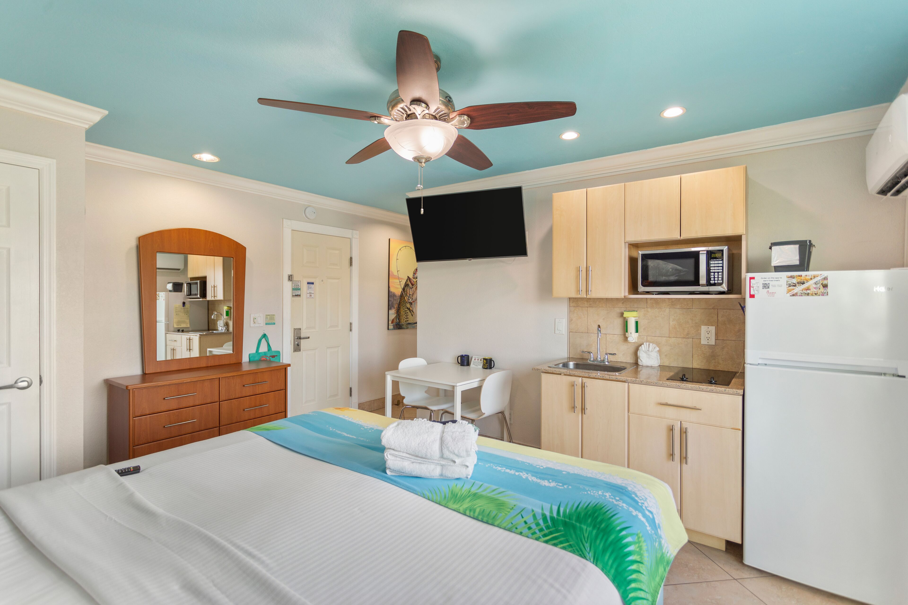A hotel room with a bed, a desk, a chair, a kitchenette, a ceiling fan, and a TV.