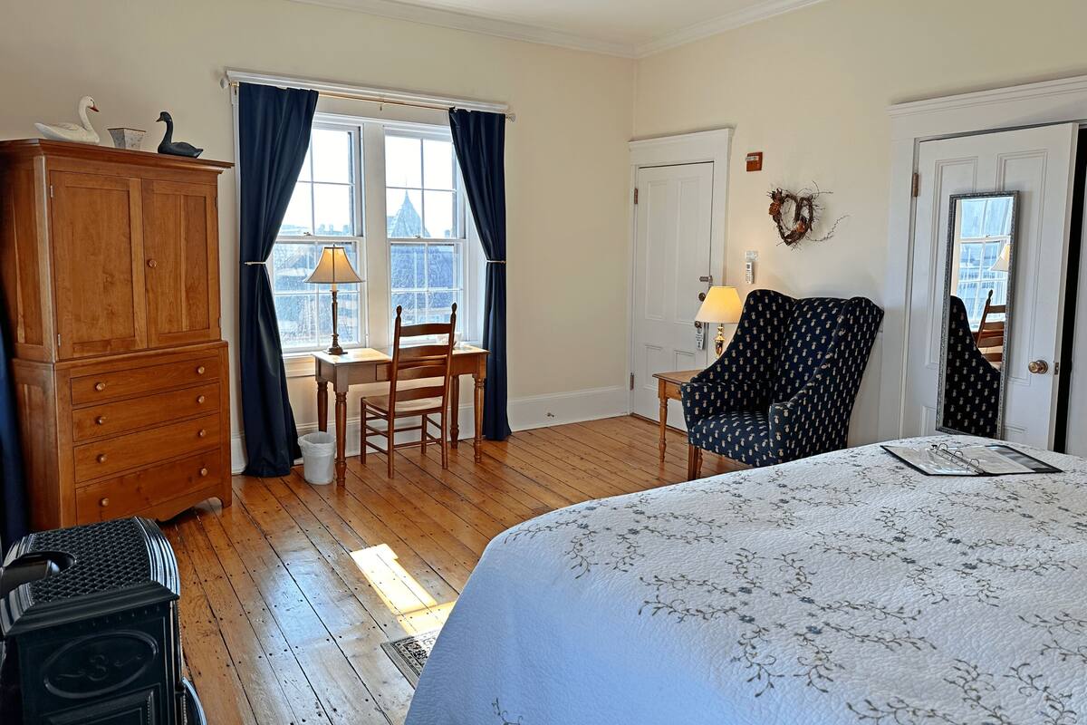 A bedroom with a wooden dresser, a desk with a chair, a patterned armchair, and a bed with a floral bedspread.