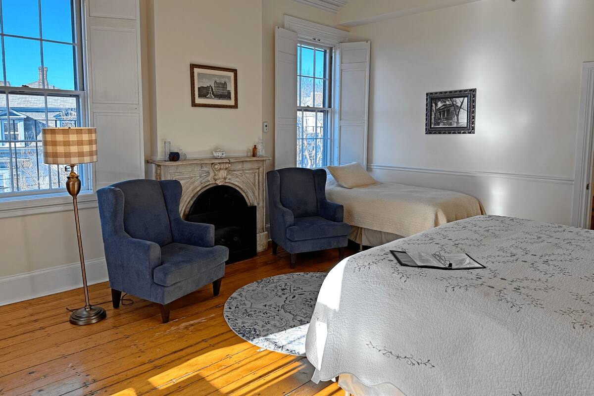 A room with a fireplace, two armchairs, a bed, and a view of the city through large windows.