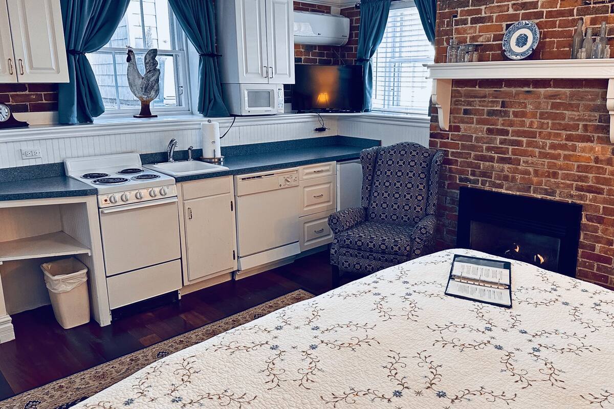 A compact kitchen with a stove, sink, and microwave, a patterned armchair, and a fireplace.