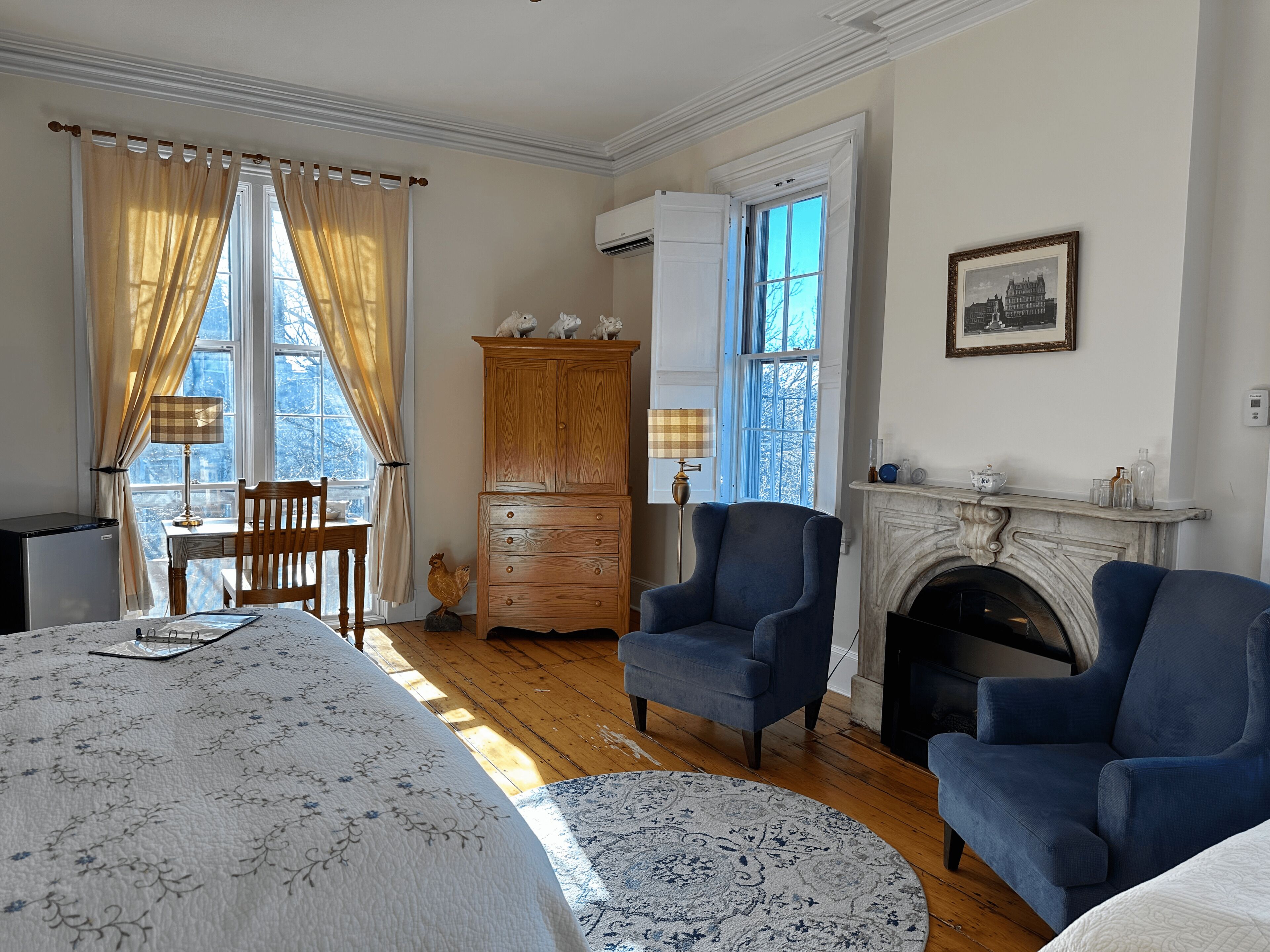 A room with a bed, two armchairs, a wooden cabinet, a fireplace, and a table with a lamp.