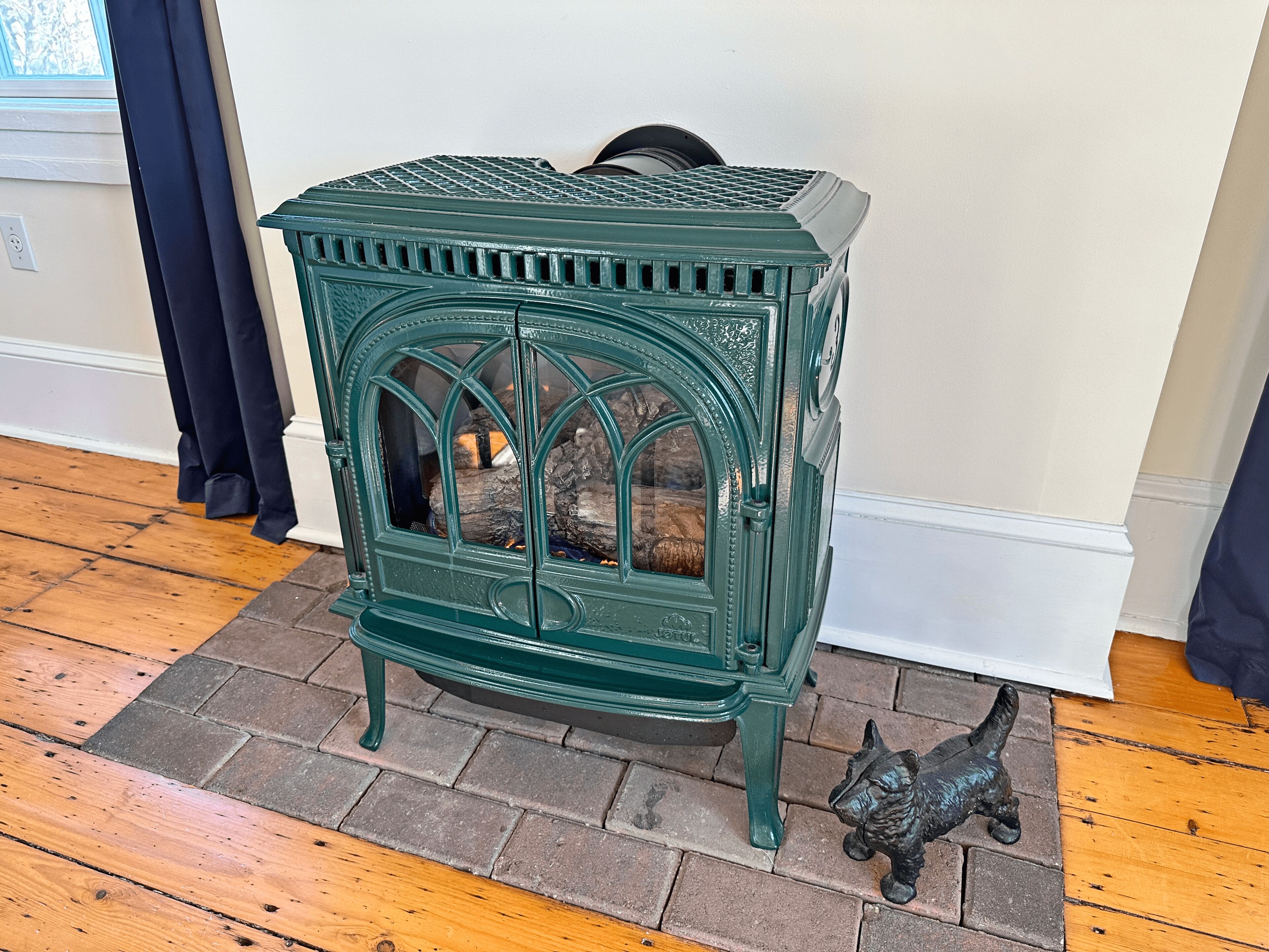 A green cast iron stove with a glass door, placed on a wooden floor with a brick border.