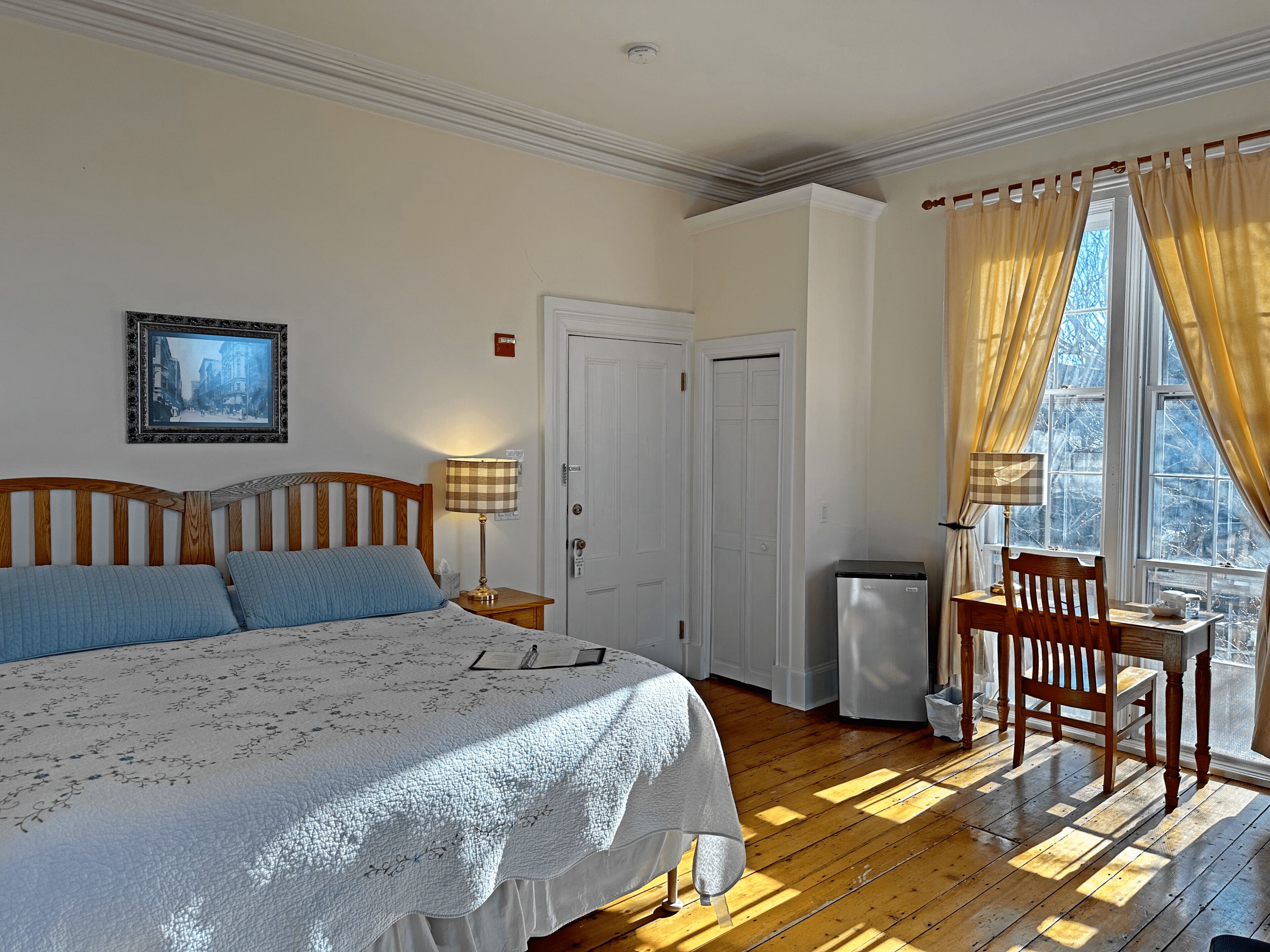 Ein Schlafzimmer mit einem großen Bett, einem Schreibtisch, einem Stuhl, einem Fenster mit Vorhängen, einer Tür und einem gerahmten Bild an der Wand.