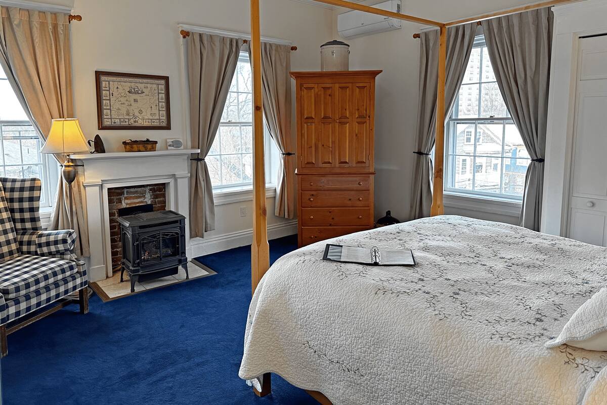 A four-poster bed with a quilt, a wooden wardrobe, a fireplace, and a checkered armchair.