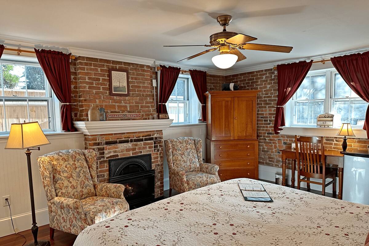 A cozy room with a fireplace, floral armchairs, a wooden desk, and a ceiling fan.