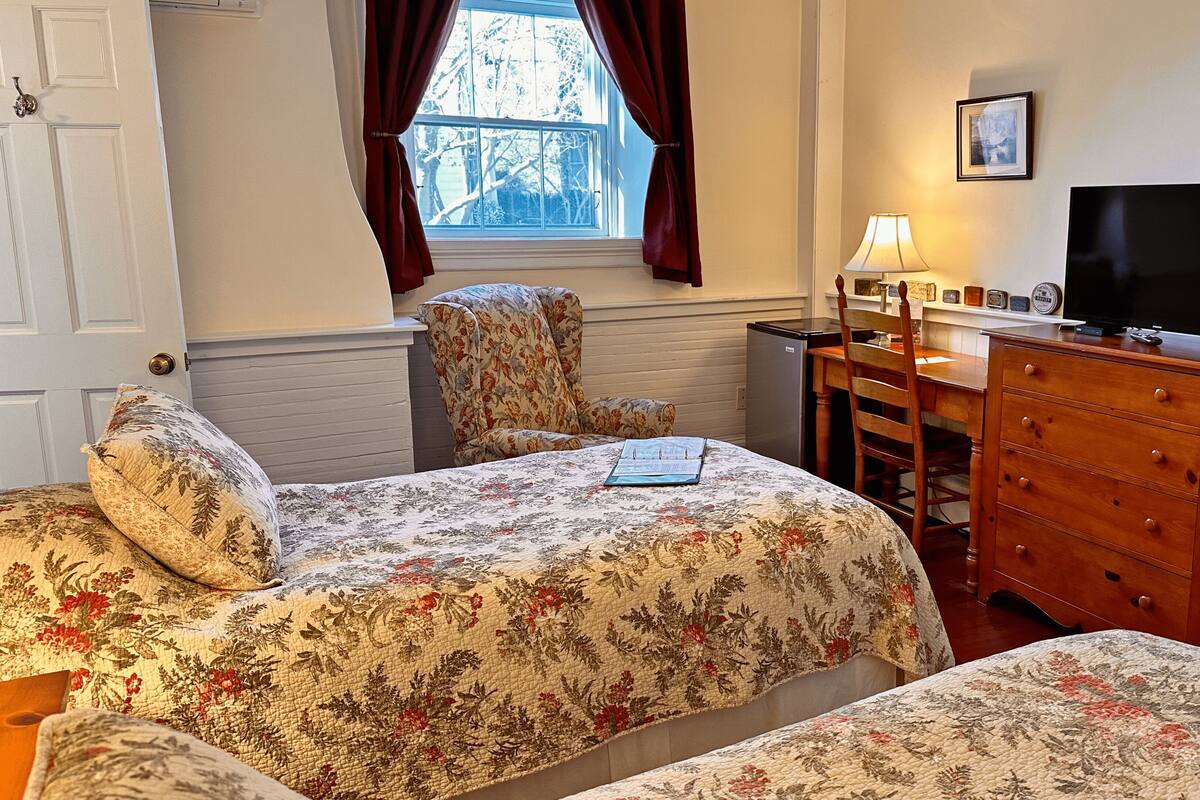 A bedroom with two beds, a desk, a dresser, a television, and a window with curtains.
