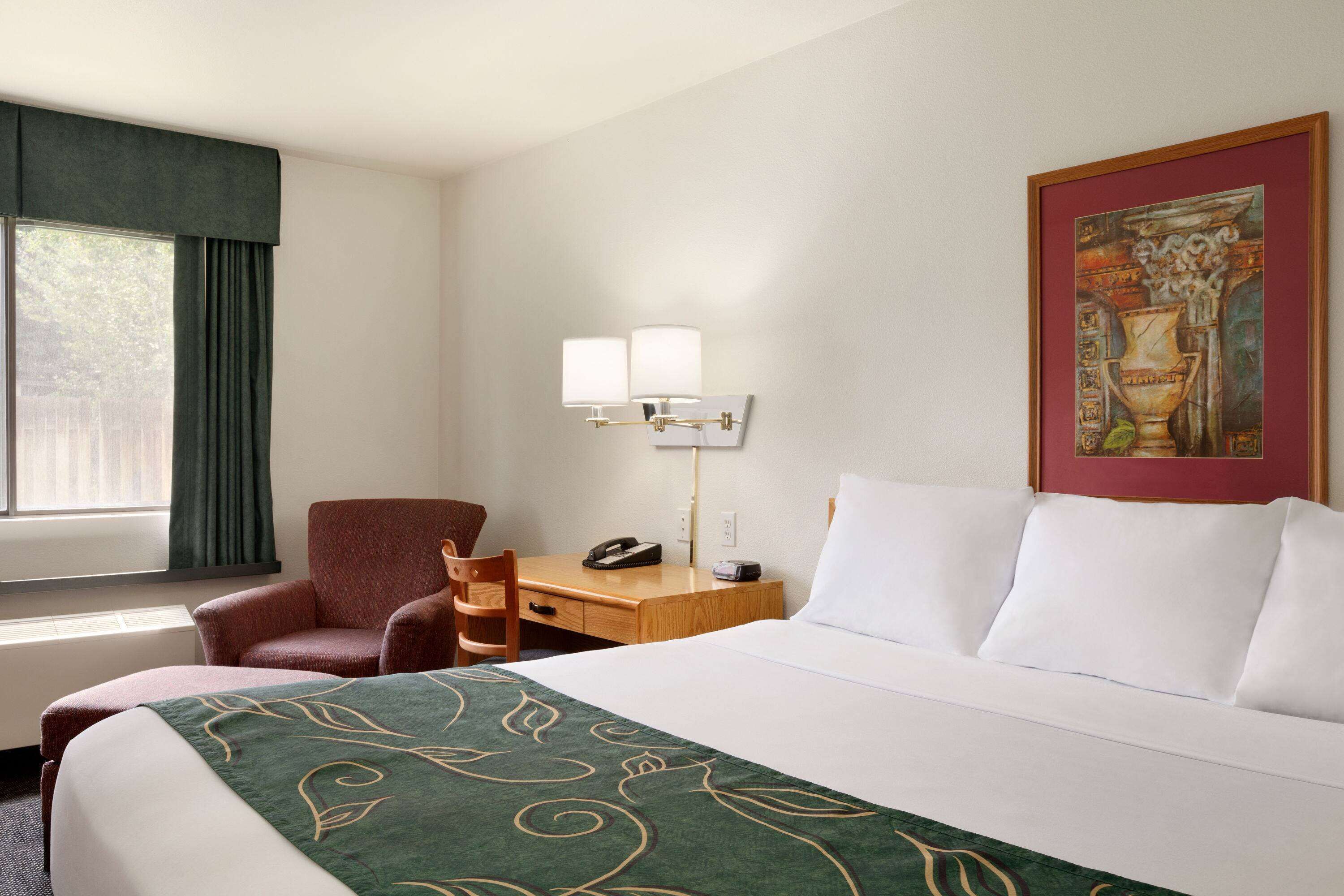 A hotel room with a bed, a desk, a chair, a lamp, and a painting on the wall.
