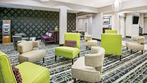 A hotel lobby with multiple seating areas, including green and beige armchairs, a patterned carpet, and a reception desk.