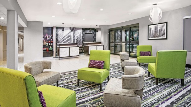 A modern lobby with green and grey chairs, a patterned carpet, and a reception desk with a cityscape photo.