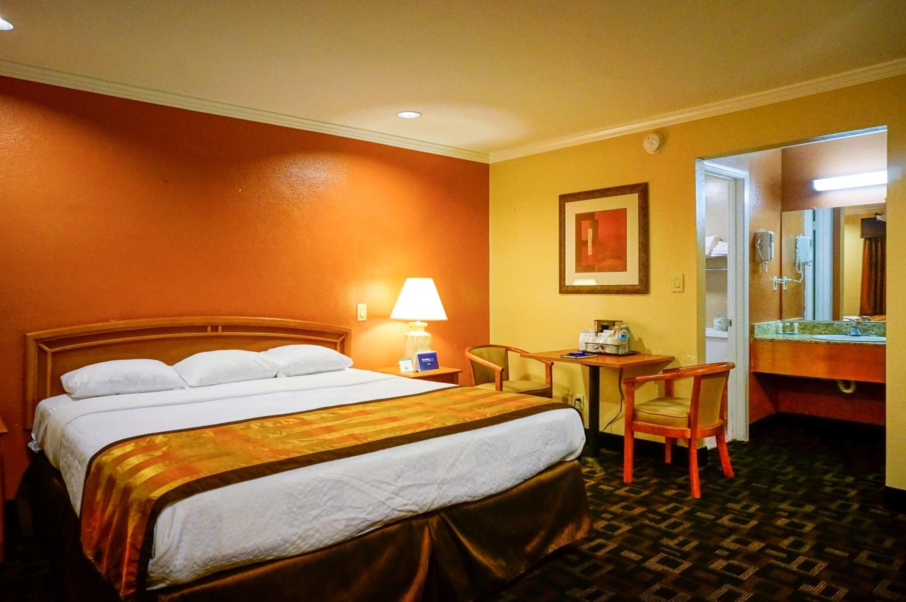 A hotel room with a large bed, a desk, a chair, a lamp, a mirror, and a bathroom visible through an open door.