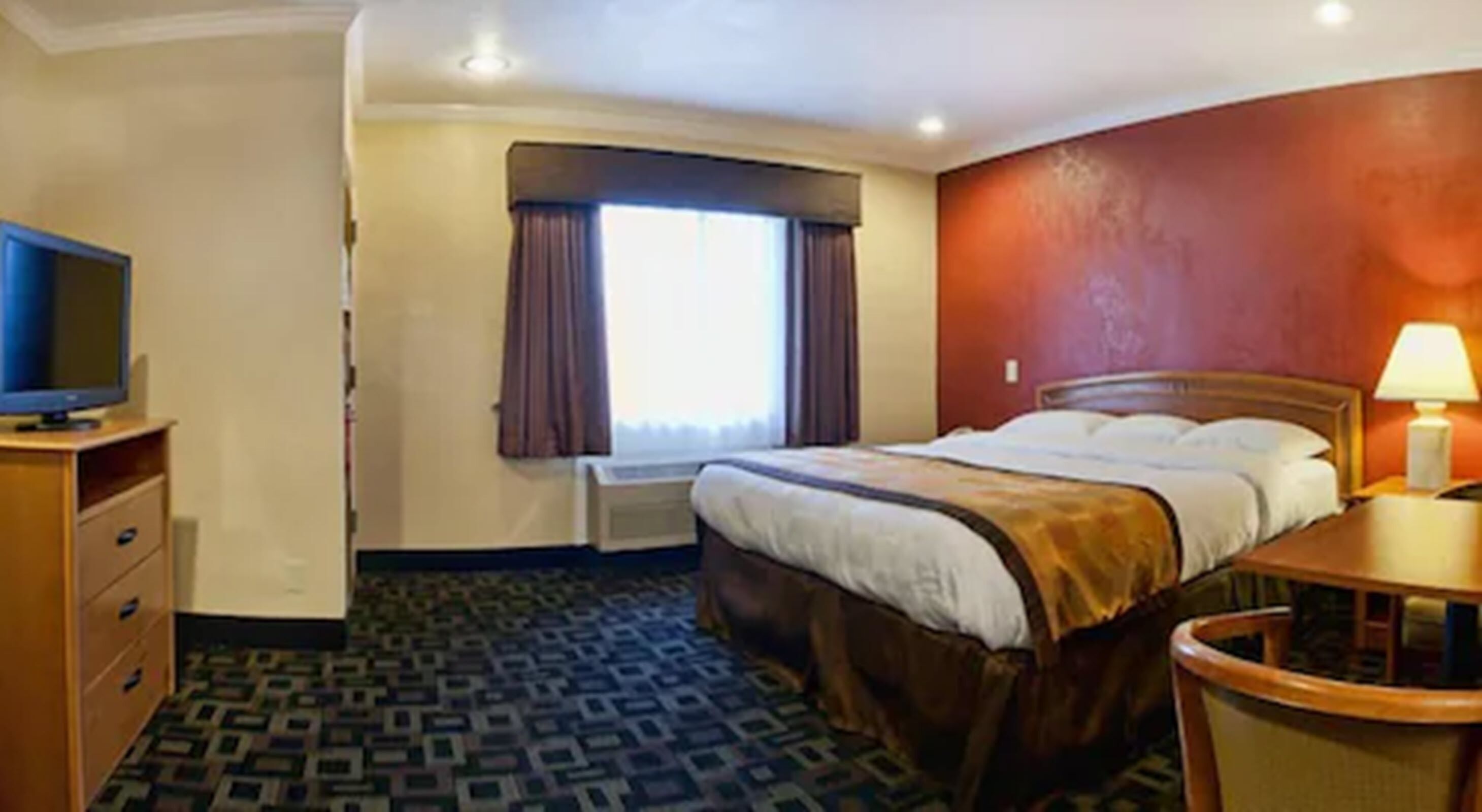 Una camera d'albergo con un letto, una scrivania con una lampada, una televisione su un comò e una finestra con tende.