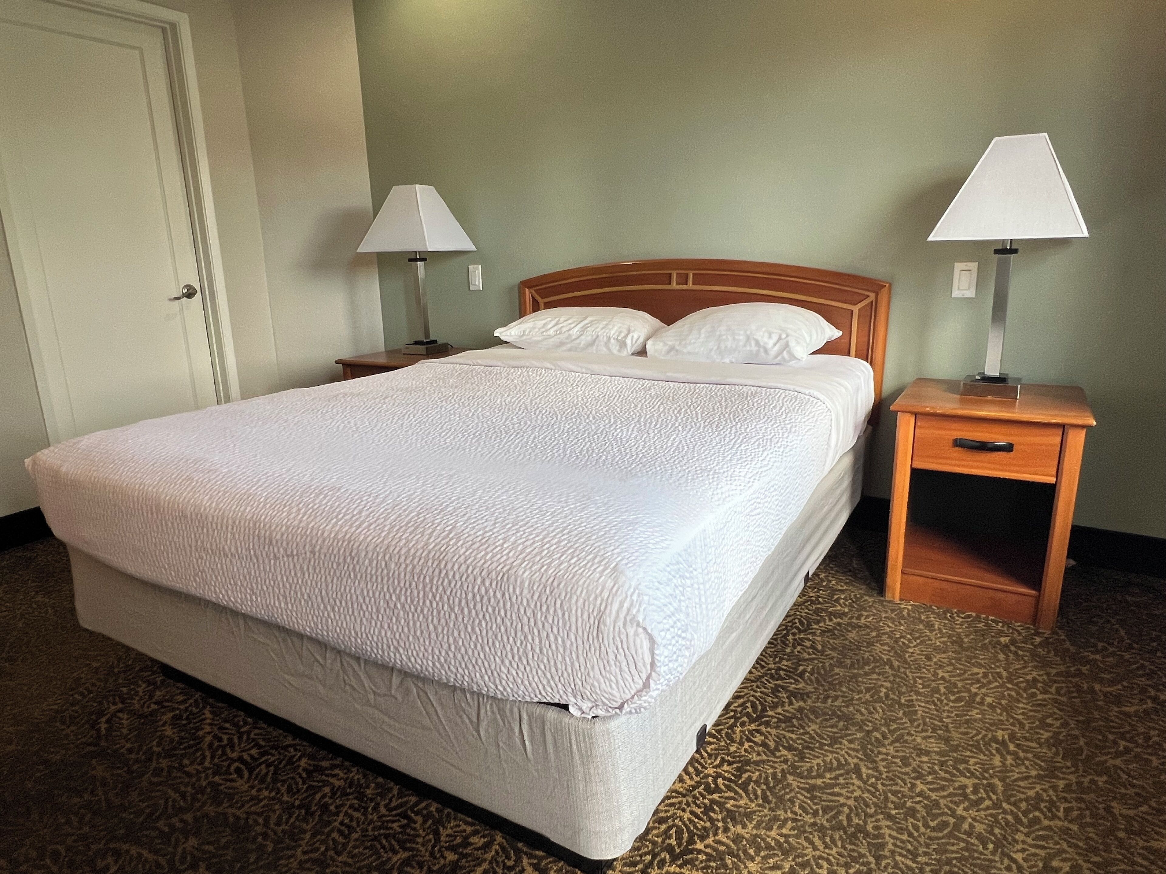 A hotel room with a large bed, two bedside tables with lamps, a wooden headboard, and a carpeted floor.