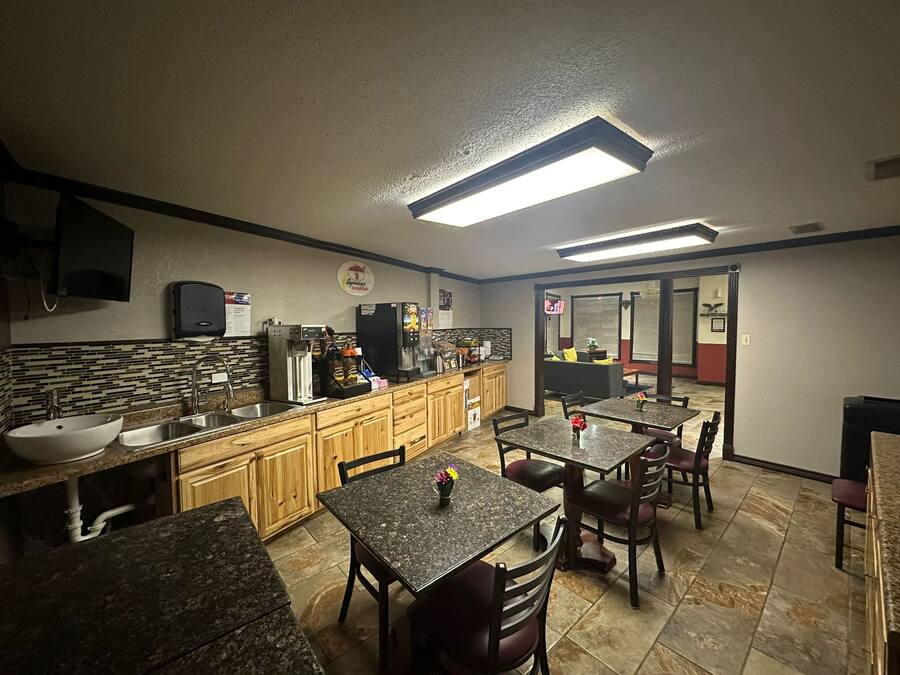 A kitchen with granite countertops, wooden cabinets, and a breakfast bar. There are two tables with chairs, a refrigerator, and a coffee machine.