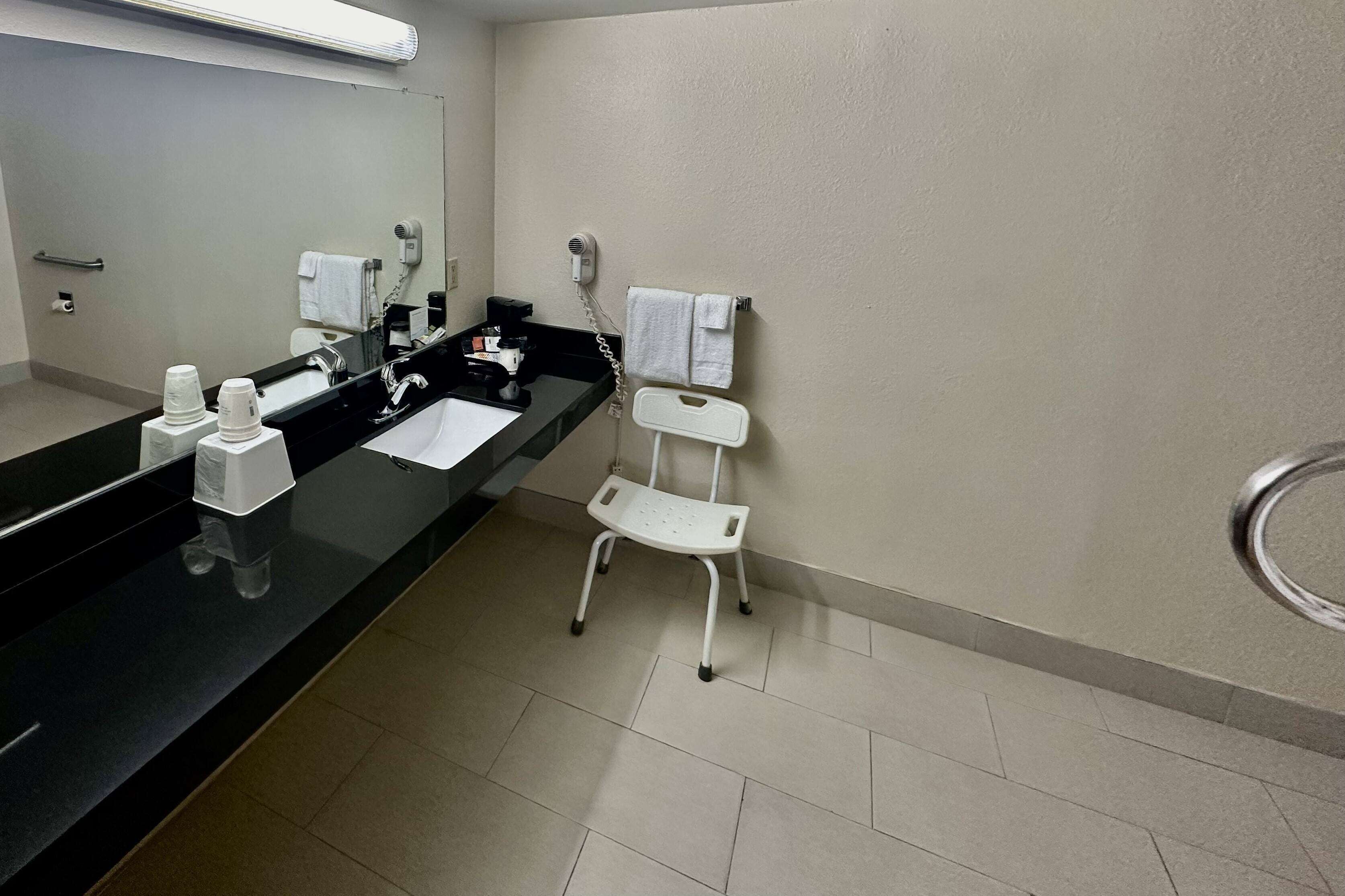 Un baño moderno con encimera negra, un lavabo blanco, un espejo, un secador de pelo y una pequeña silla blanca.