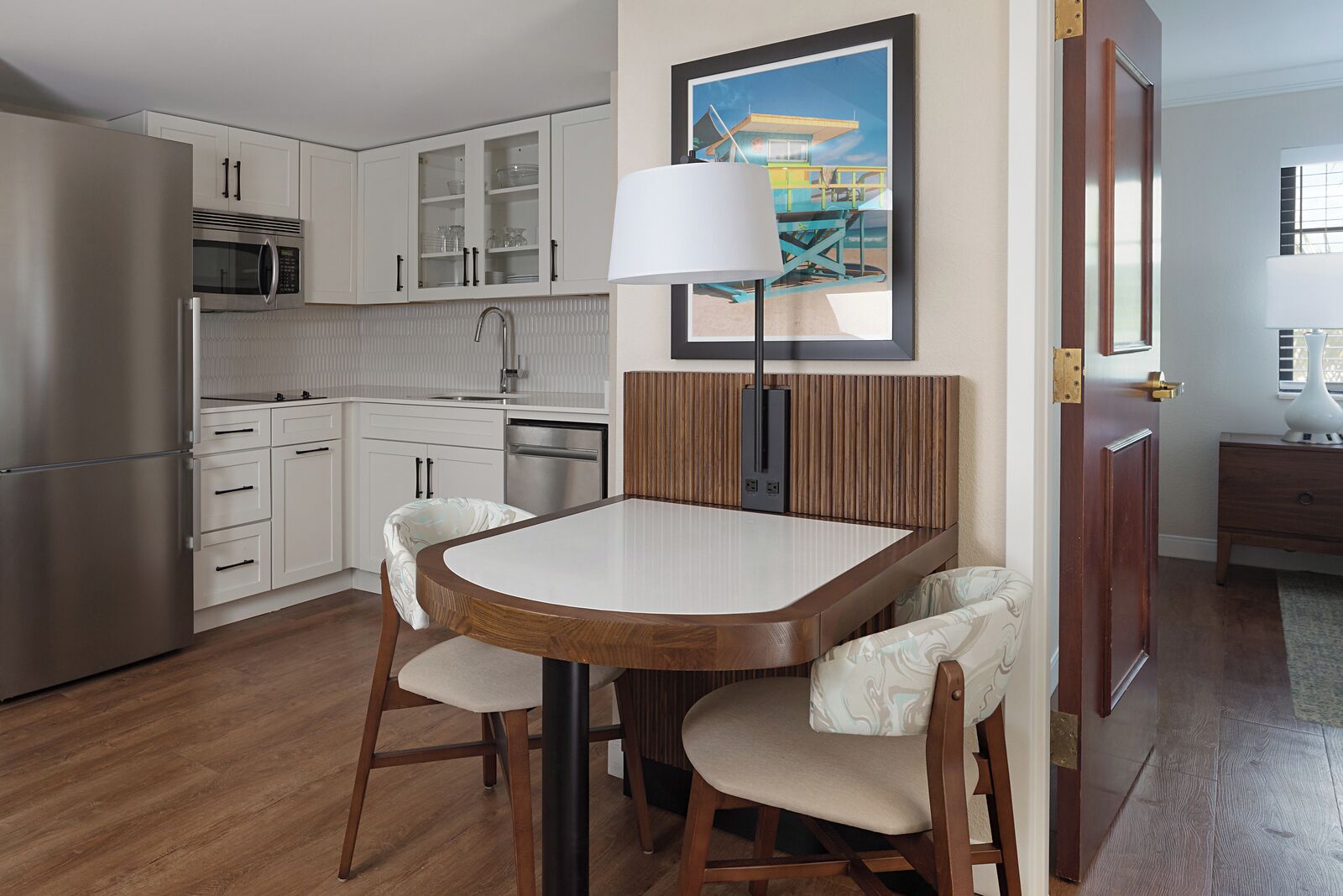 Cocina compacta con alacenas blancas, microondas y un lavabo. Hay una mesa de comedor con dos sillas y una lámpara.