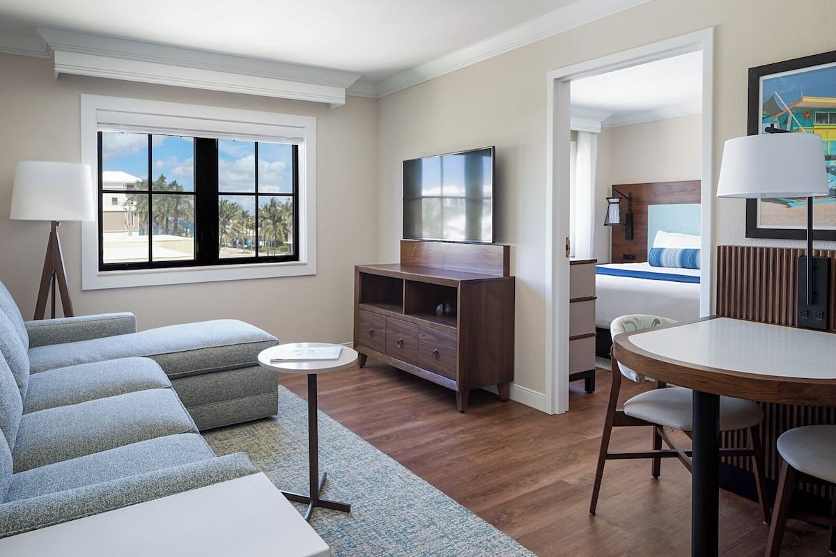 Una habitación de hotel moderna con sofá, una mesita redonda pequeña, televisor y cama con colcha a rayas azules y blancas.