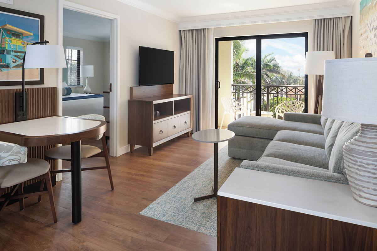 Habitación de hotel con zona de comedor, sofá, televisión y balcón con vista a palmeras.
