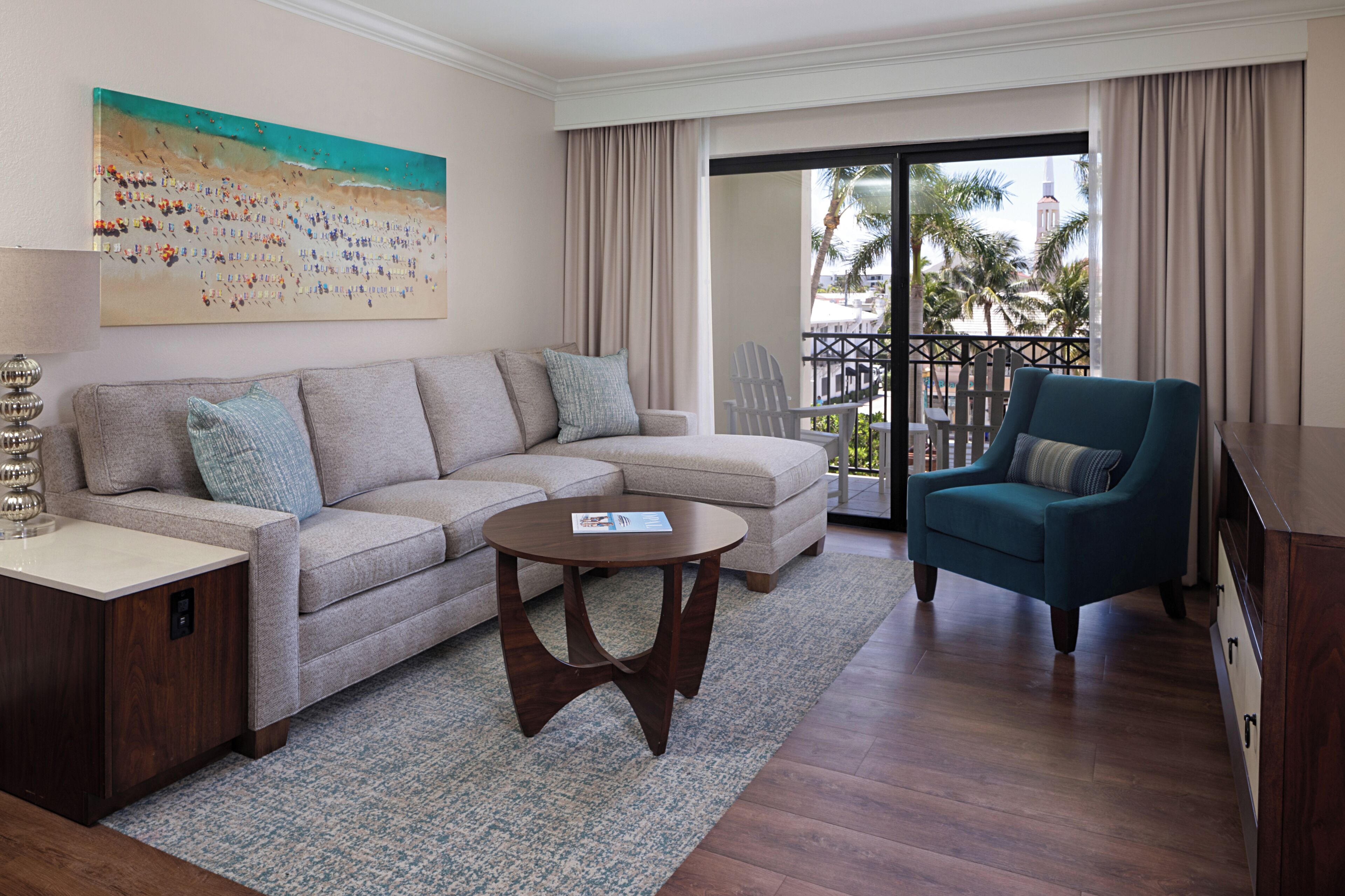 A living room with a sofa, a teal armchair, a coffee table, and a painting of a beach scene.