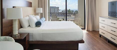 A hotel room with a large bed, a TV, a desk with a lamp, and a balcony with chairs.