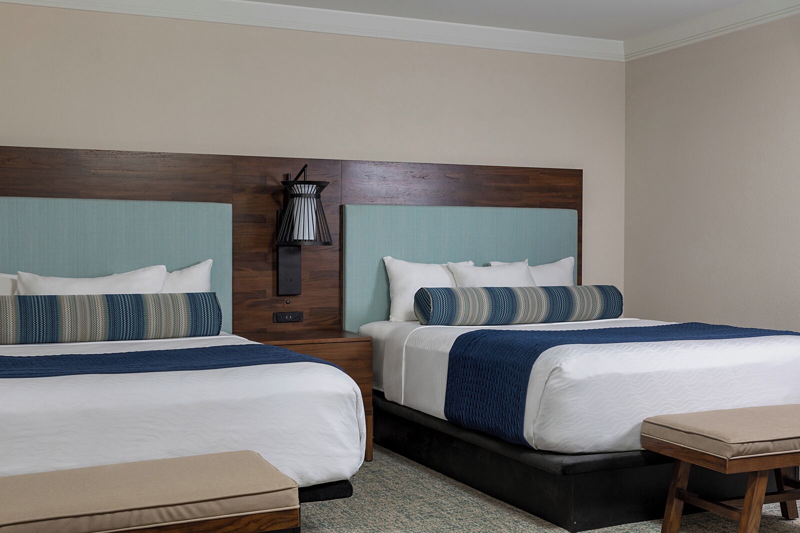 A hotel room with two beds, a nightstand, a lamp, and a small table.