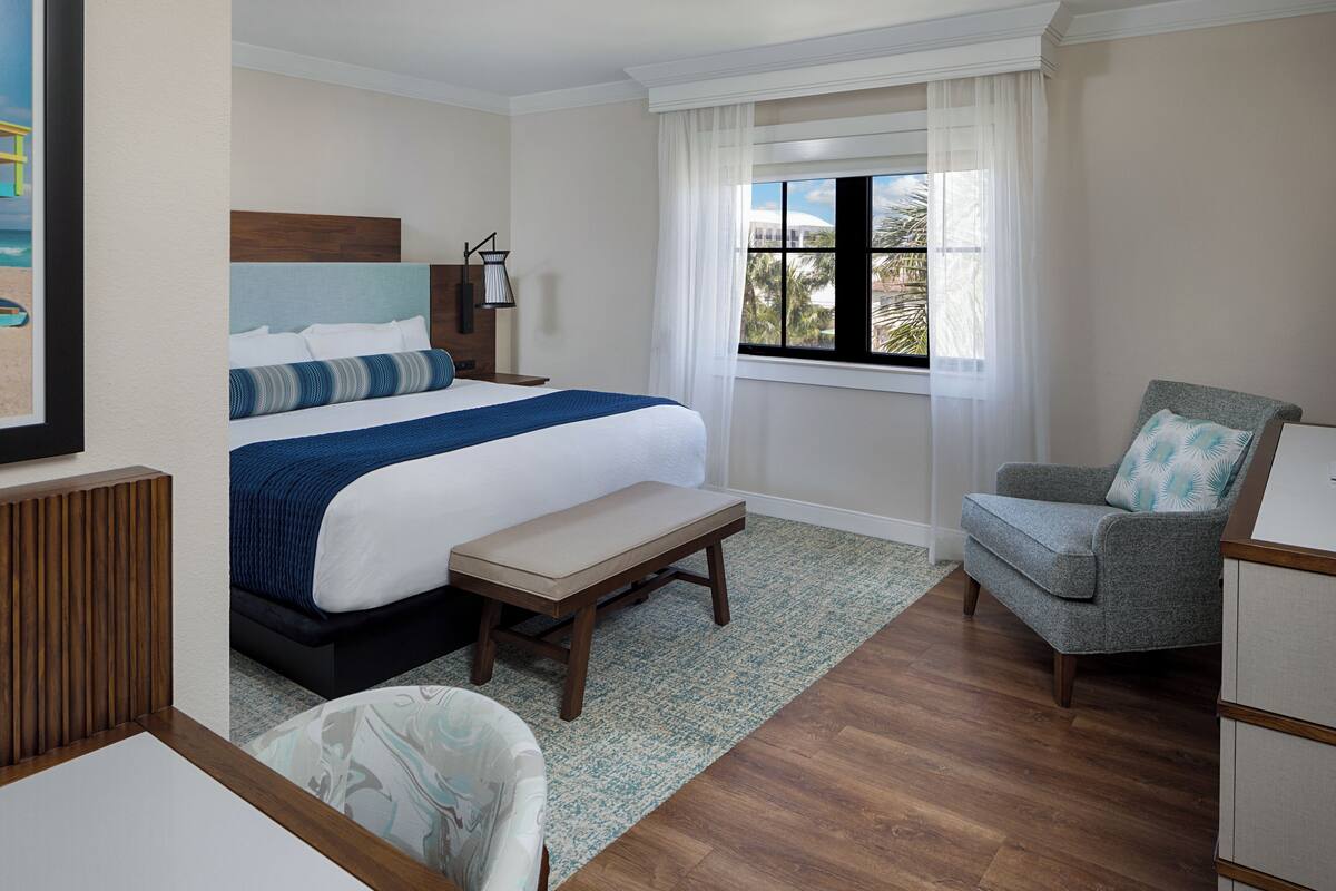Habitación de hotel con cama, una silla, un escritorio, una ventana con cortinas y vista a palmeras.