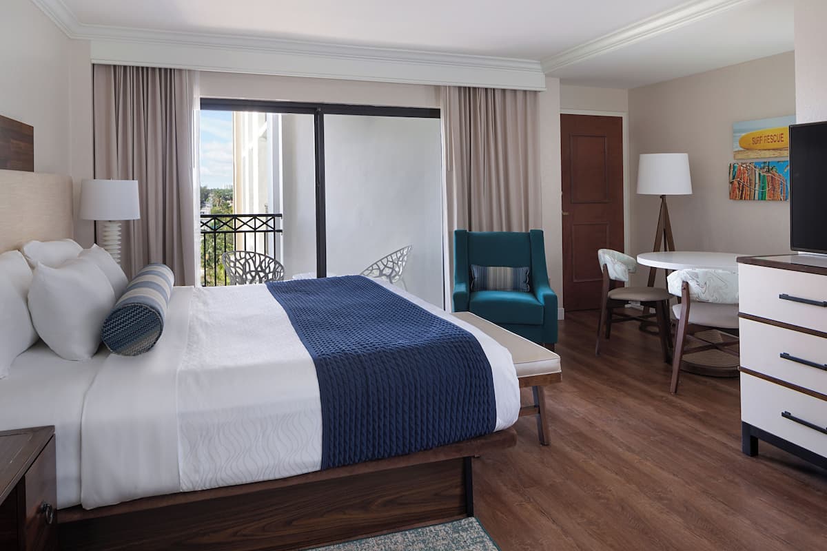 Habitación de hotel con una cama grande, un escritorio, una silla, un televisor y un balcón con cortinas.