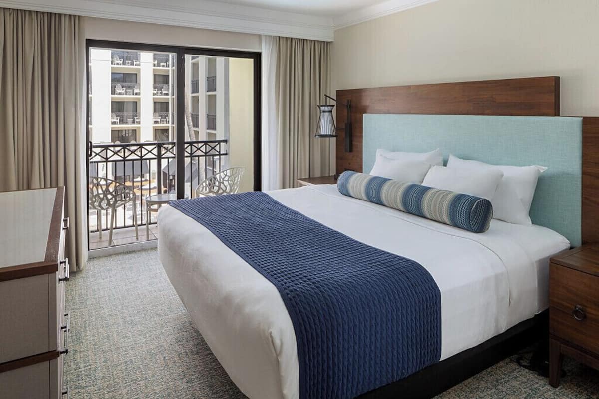 Habitación de hotel con una cama grande, cabecera de madera, balcón con vistas al edificio y un escritorio con silla.