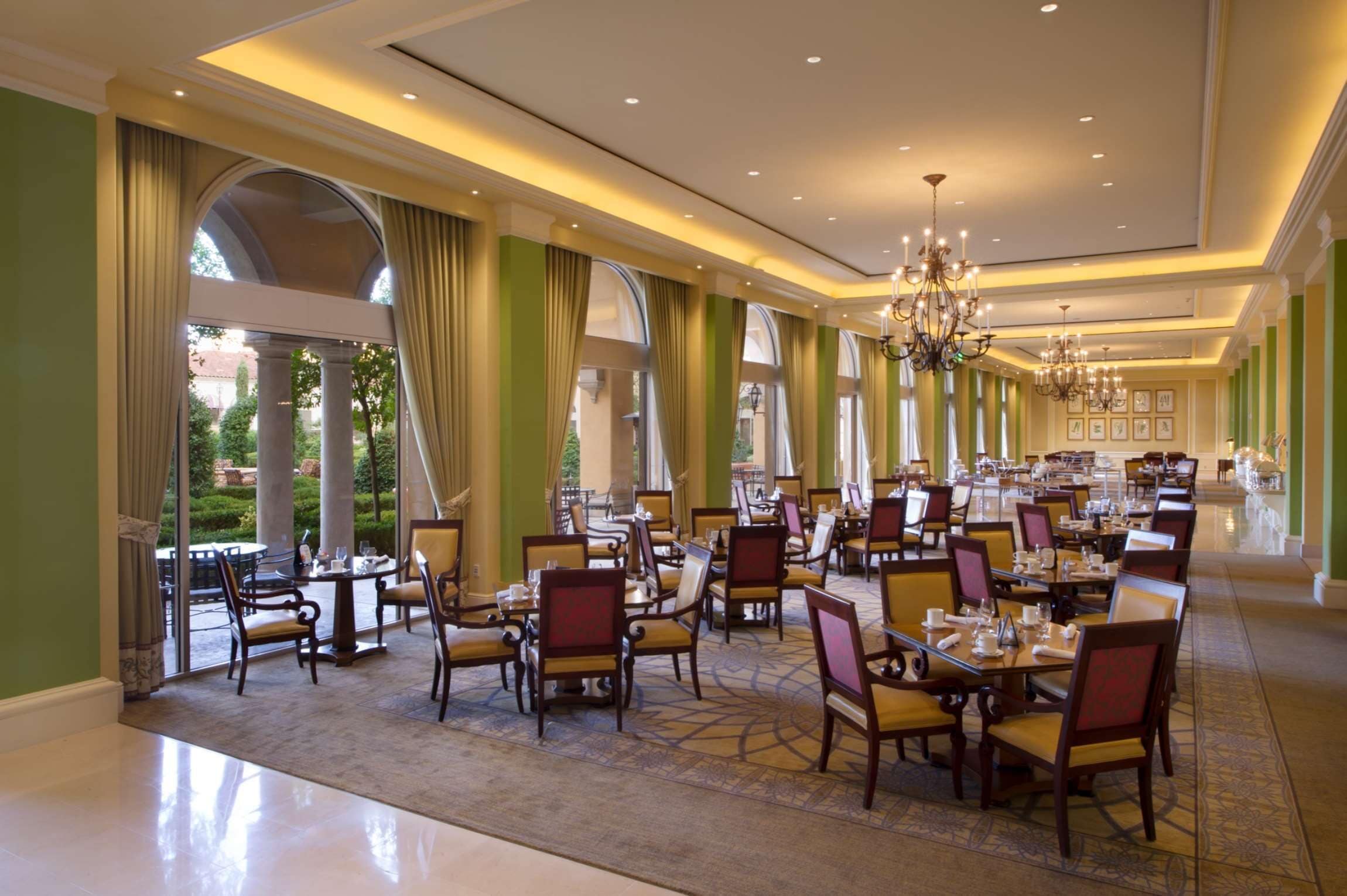 A spacious dining area with multiple tables and chairs, large windows with curtains, and a chandelier.