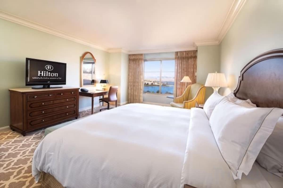 A hotel room with a large bed, a desk with a chair, a television, and a view of the outdoors.