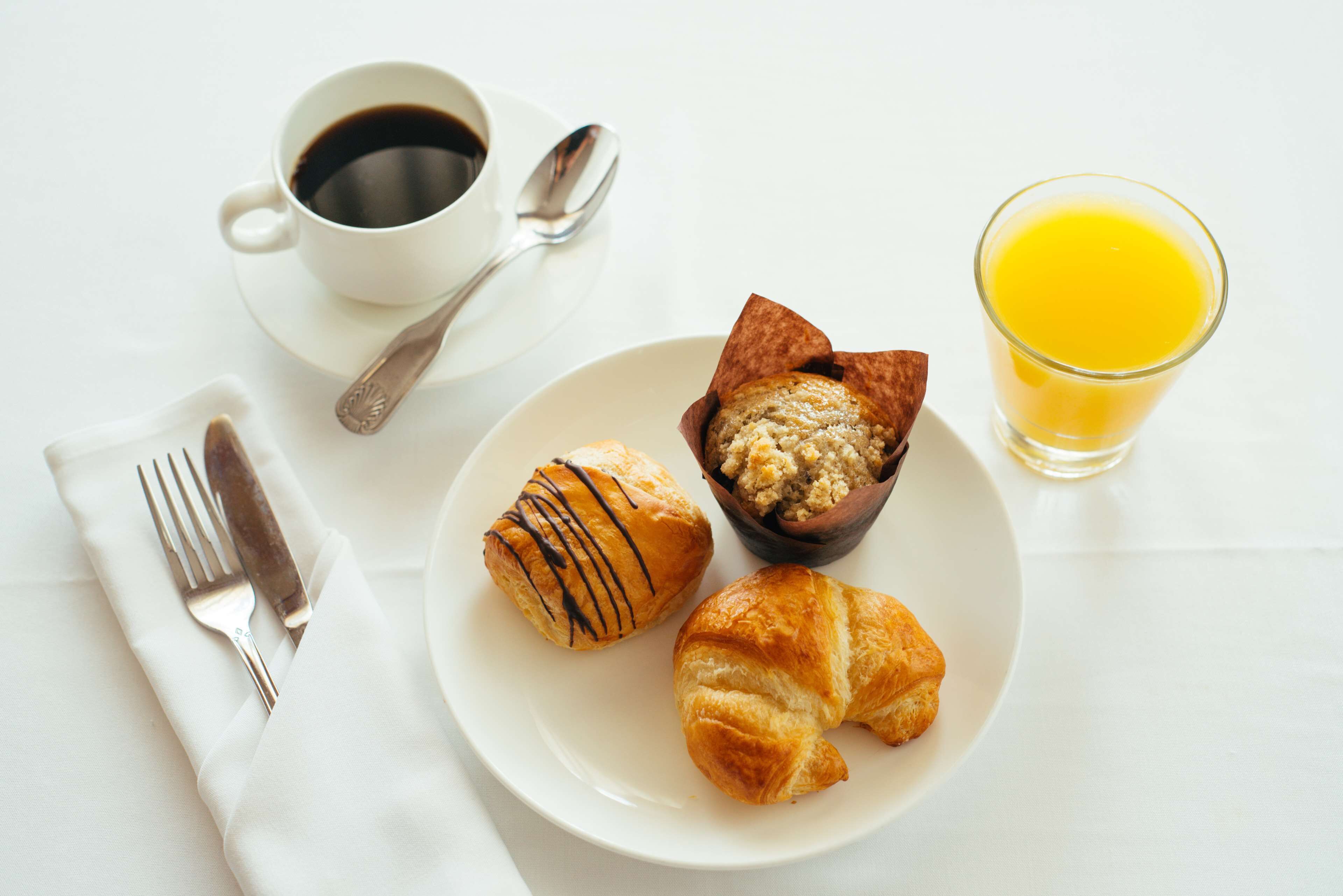 A breakfast setting with a plate of croissants, a muffin, a cup of coffee, a glass of orange juice, and cutlery on a white table.