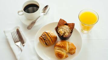 A breakfast setting with a plate of croissants, a muffin, a cup of coffee, a glass of orange juice, and cutlery on a white table.