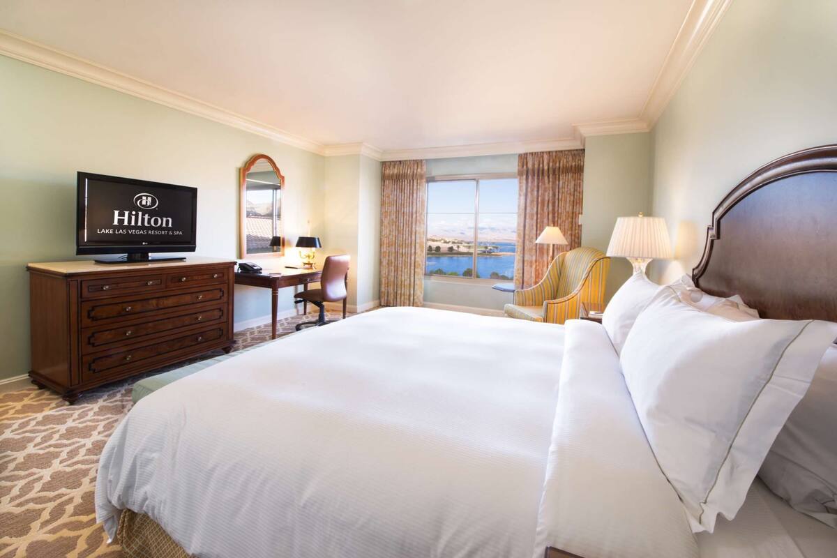 Een hotelkamer met een groot bed, een kaptafel, een bureau, een televisie waarop het Hilton-logo staat, een raam met gordijnen en uitzicht op een waterpartij.