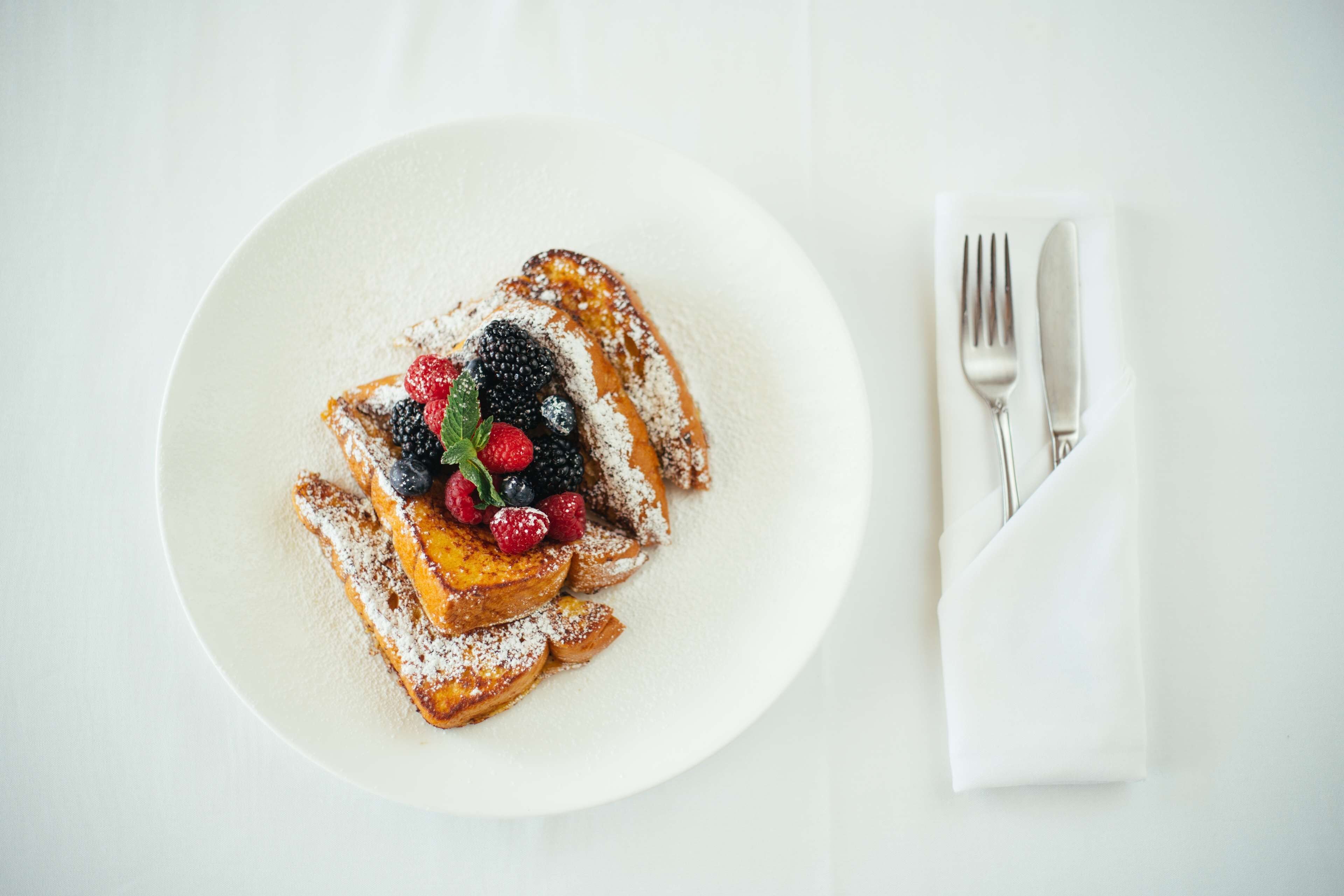 A plate of French toast with berries and powdered sugar, accompanied by a fork and knife on a napkin.