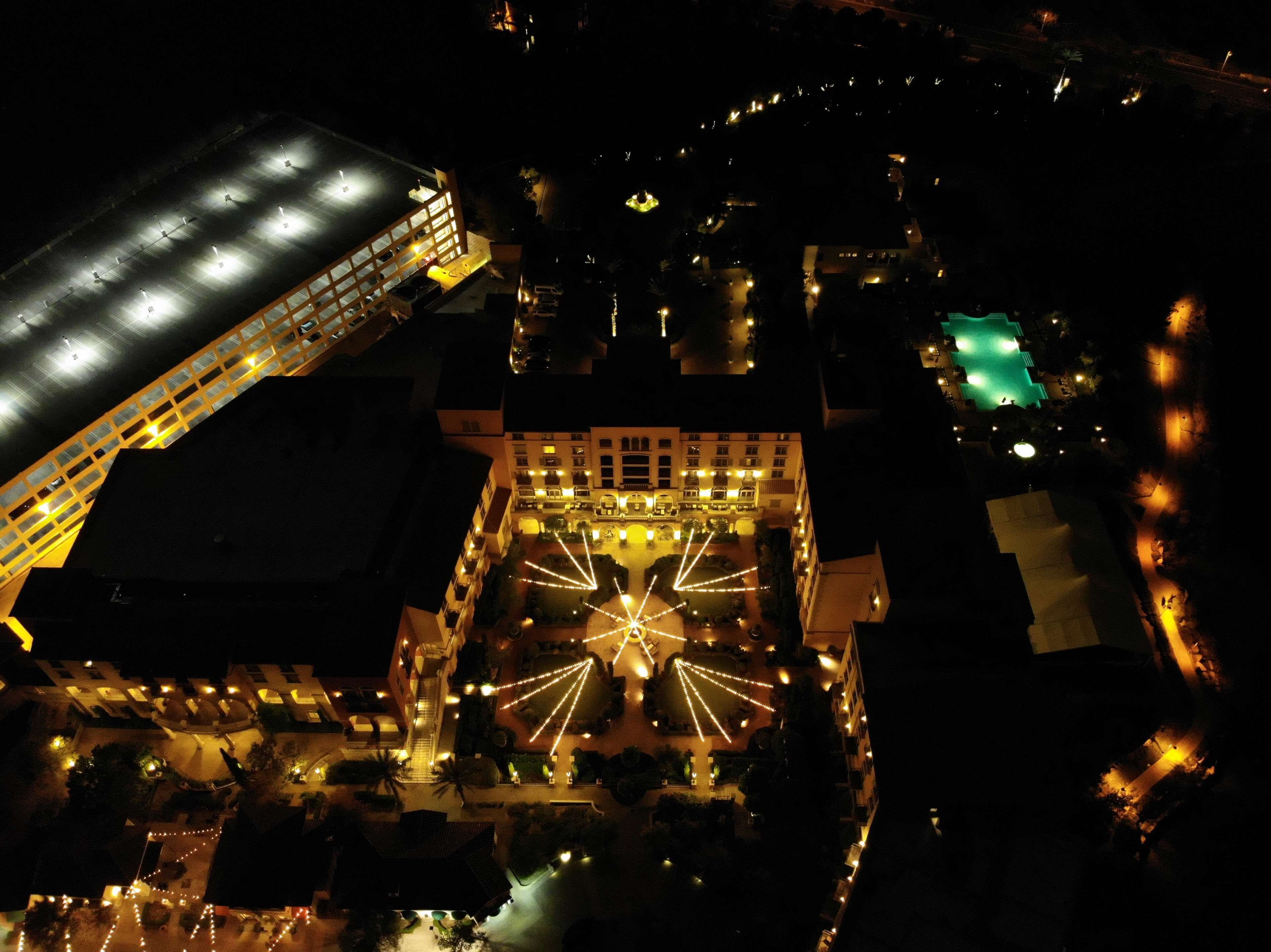 Una vista aerea di un hotel con un cortile centrale illuminato da luci a tenda, circondato da edifici con balconi e una struttura verdeggiante a destra.