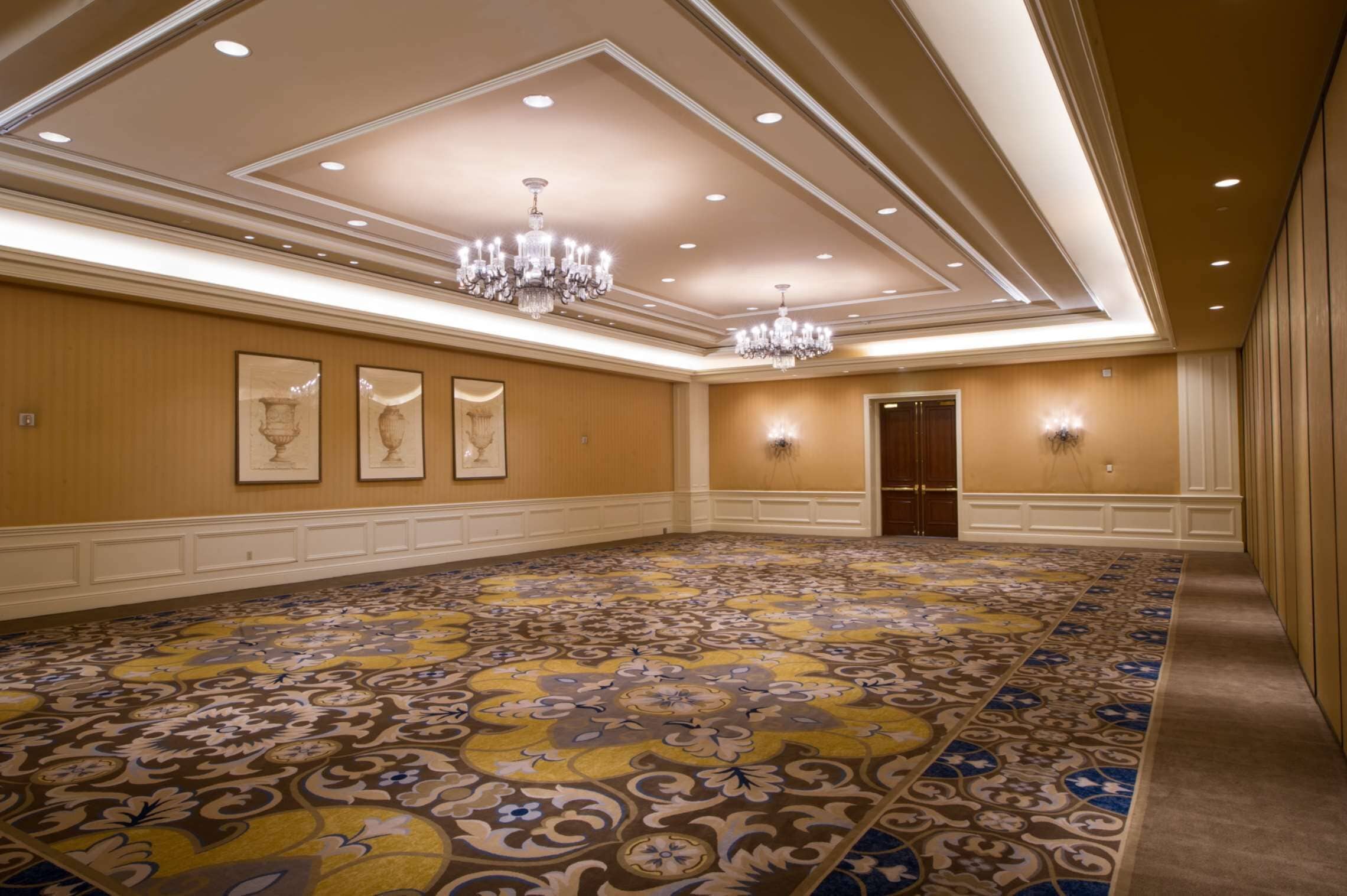 A large conference room with a patterned carpet, chandeliers, framed artwork on the walls, and recessed lighting.