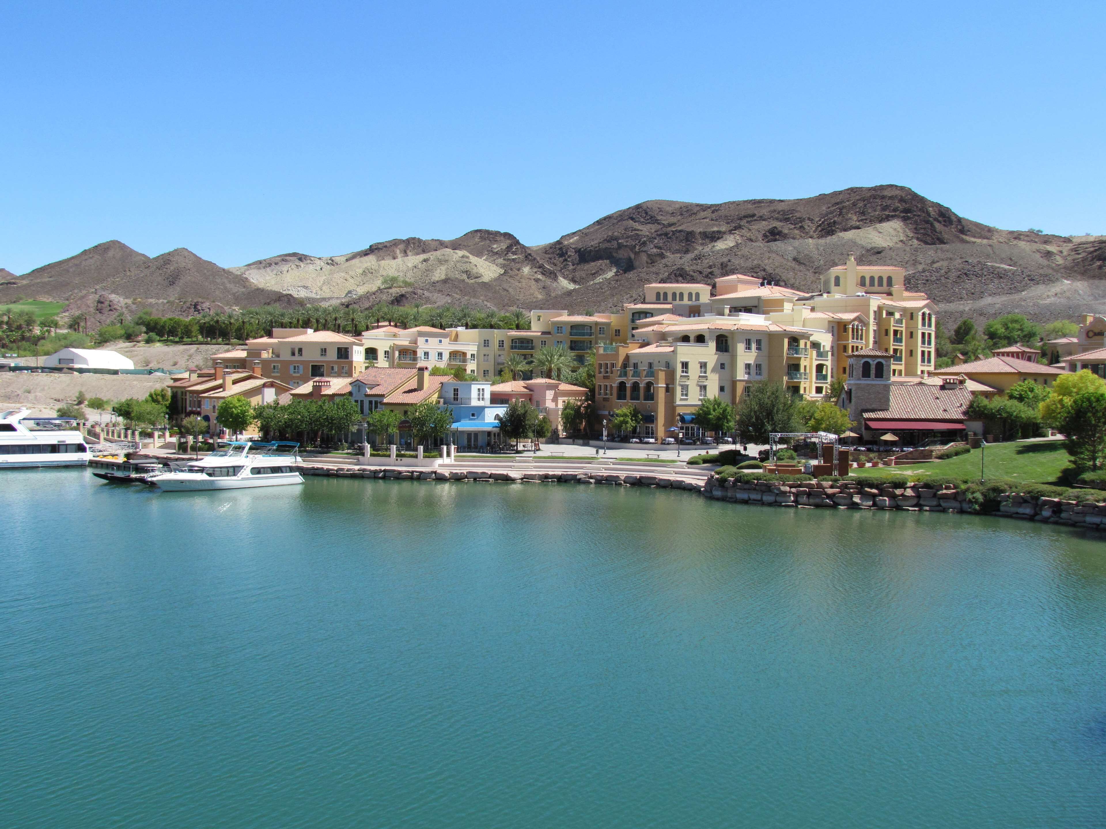 A marina with boats docked, a waterfront promenade, and a cluster of Mediterranean-style buildings with balconies.