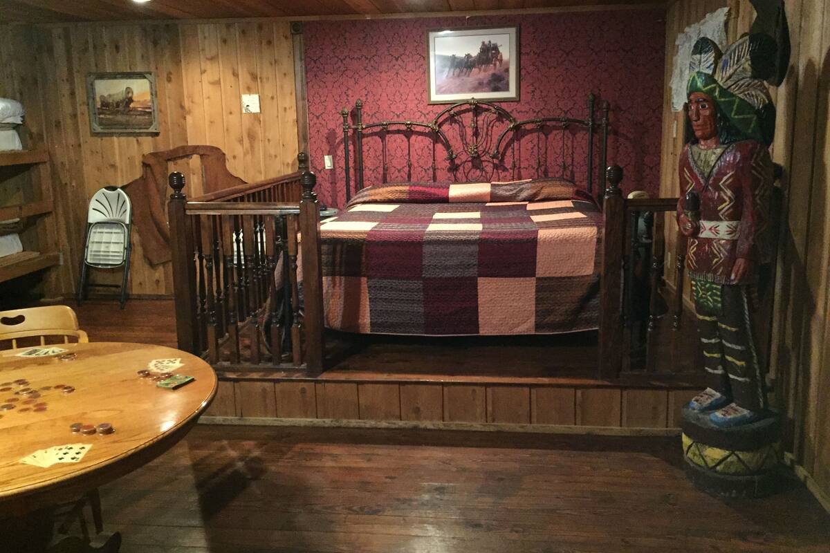 A room with a wooden floor, a bed with a checkered blanket, a wooden chair, a small table with playing cards, and a wall with a framed picture and a statue.