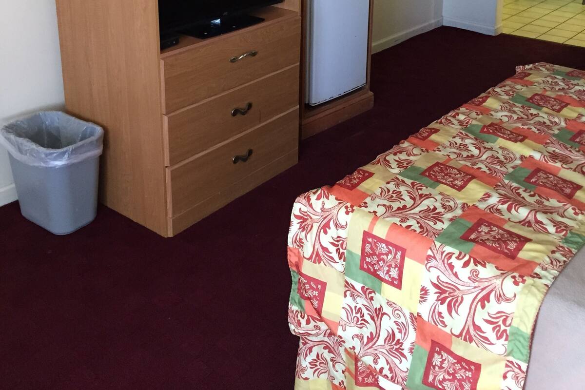 A hotel room with a wooden TV stand, a small refrigerator, a microwave, a trash bin, and a bed with a colorful quilt.
