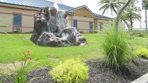 A landscaped area with a rock formation, grass, and a building with a blue roof in the background. - Wildwood Inn Tropical Dome & Theme Suites (Florence)