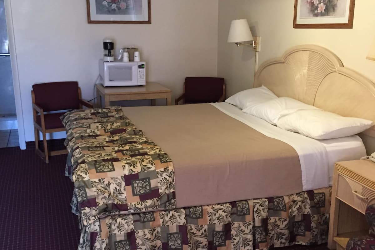 A hotel room with a bed, two chairs, a microwave, a lamp, and two framed pictures on the wall.