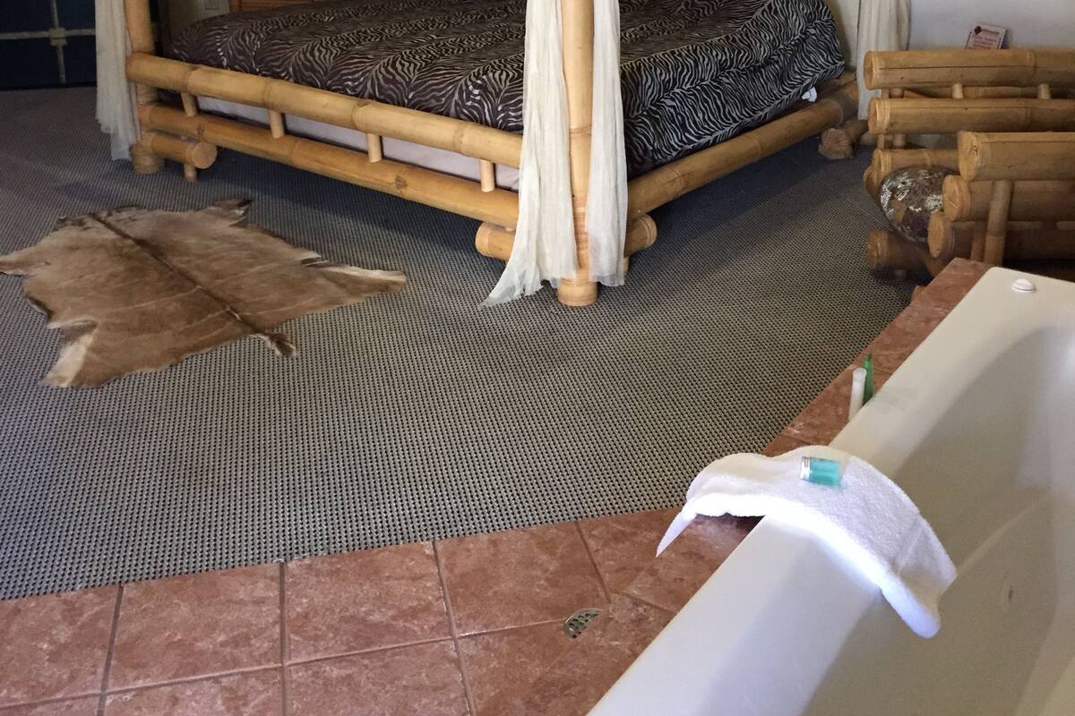 A room with a four-poster bed, a small bedside table, a large window, and a bathroom with a bathtub.