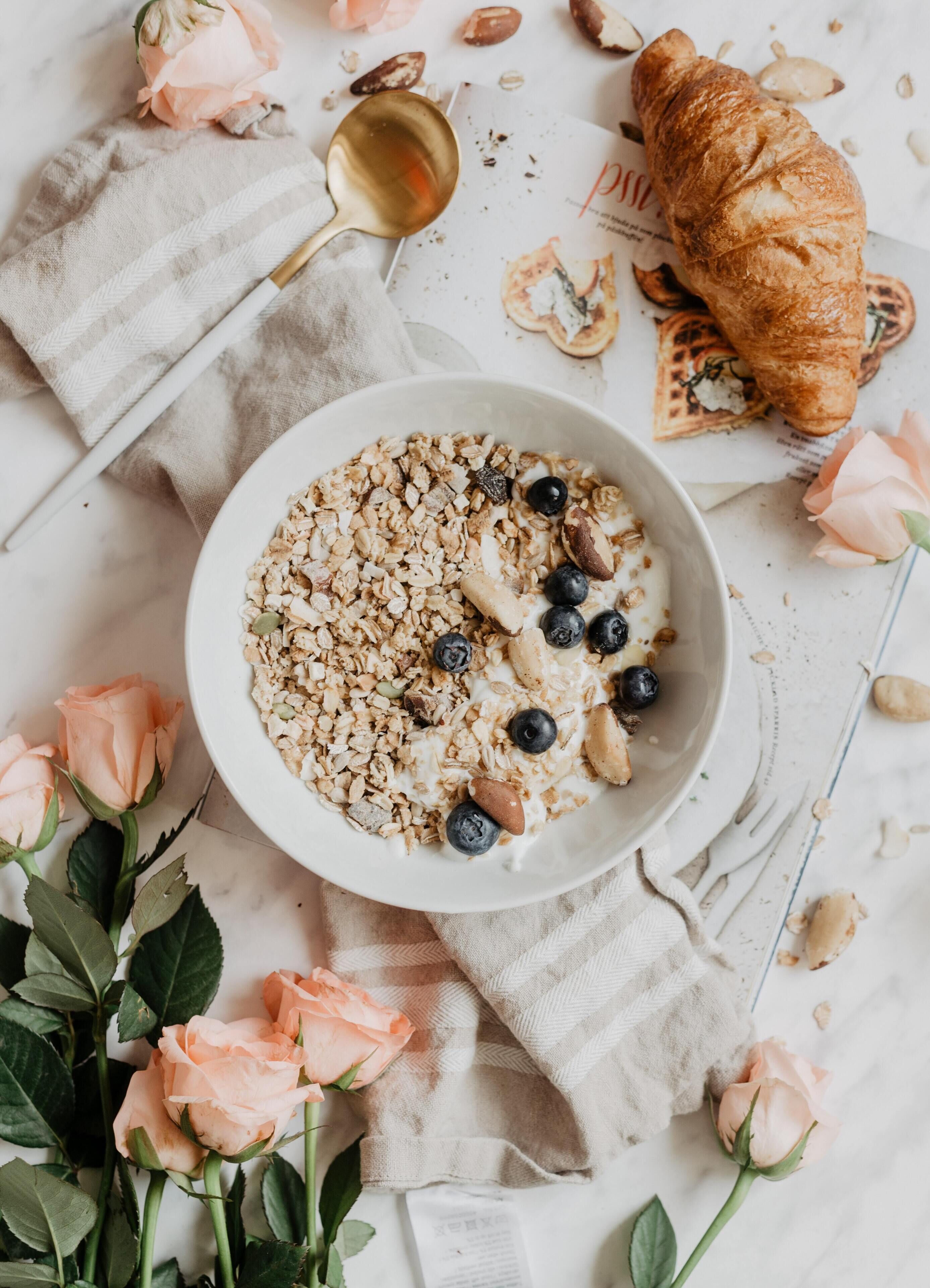 Eine Schüssel Haferflocken mit Blaubeeren und Mandeln, ein Croissant und ein Löffel auf einem Tisch mit karierter Tischdecke.