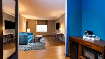 Ein modernes Wohnzimmer mit einem blauen Sofa, einem Couchtisch und einem Fernseher. Es gibt eine Küchenzeile mit einer Mikrowelle und einer Kaffeemaschine.