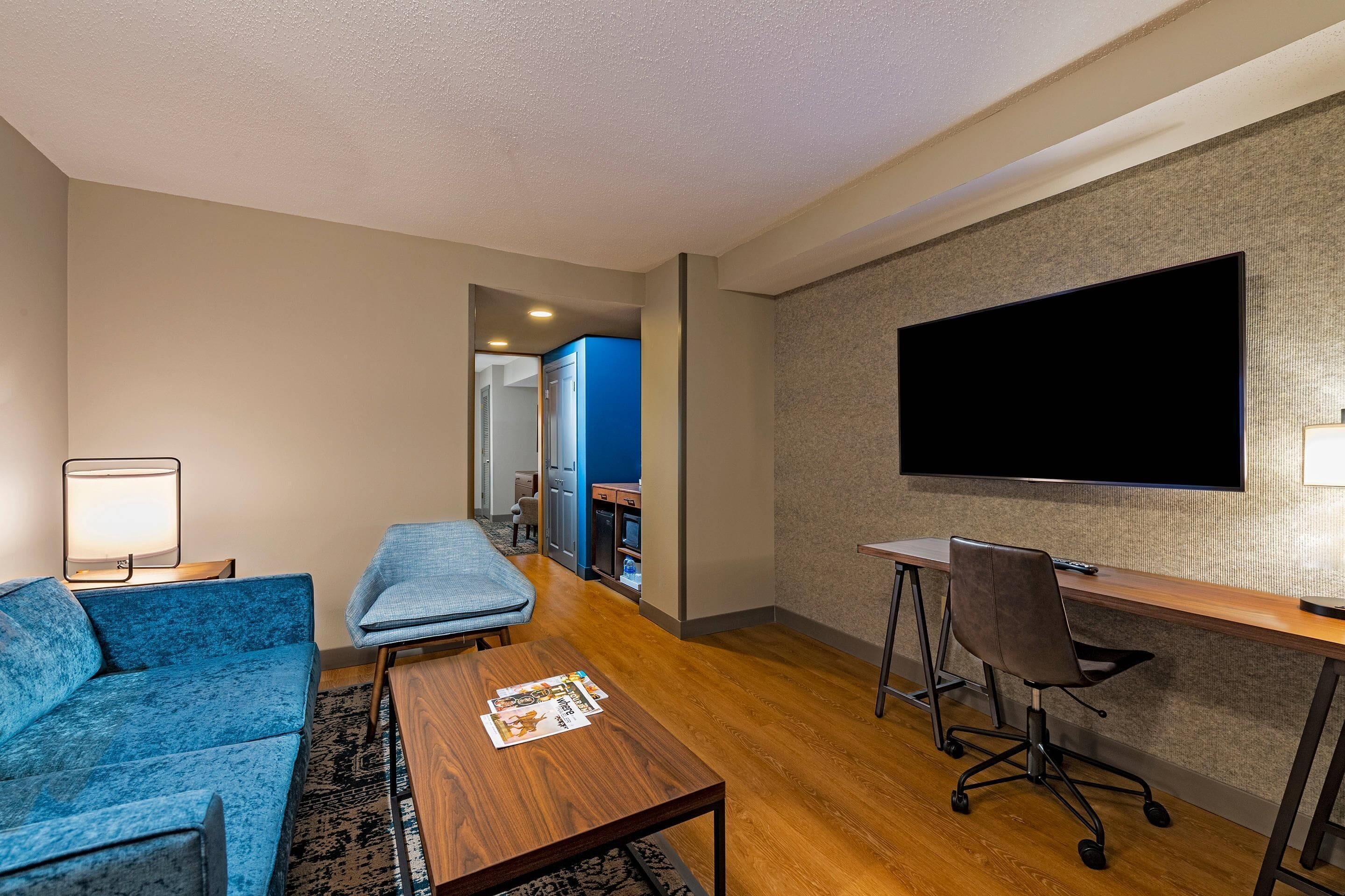 A modern hotel room with a blue sofa, a wooden coffee table, a desk with a chair, and a flat-screen TV mounted on the wall.