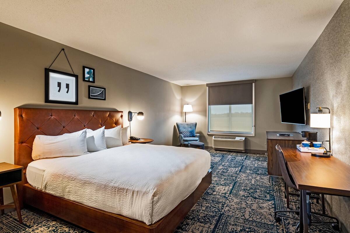 A hotel room with a large bed, a desk, a chair, a TV, and framed pictures on the wall.